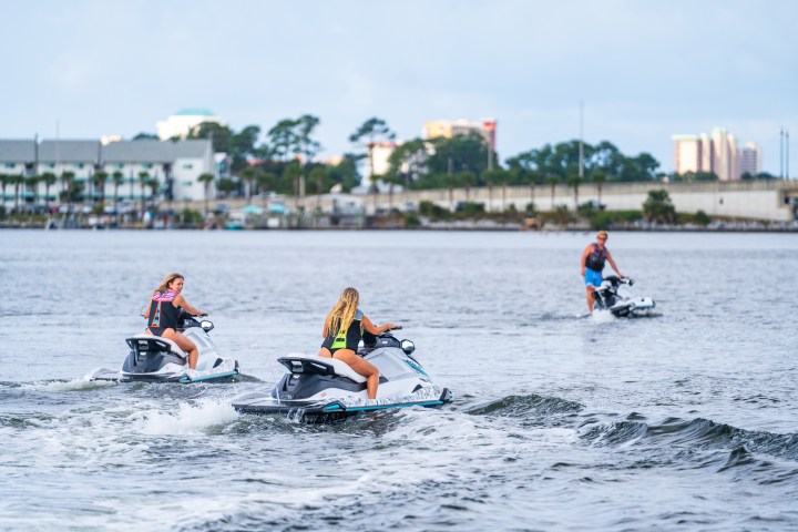 Three people riding jet skis on a calm water body with a cityscape background.