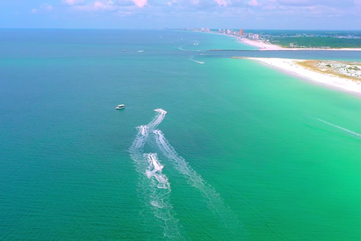 Aerial view of a boat leaving white trails on turquoise sea near a sandy coastline.