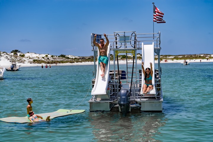 People on boat with slides, in water, child on floating mat, American flag above, sandy shore in background.
