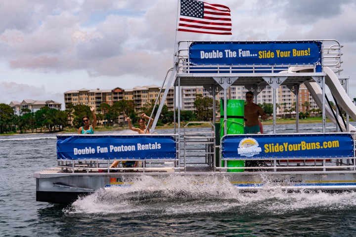 Pontoon boat with slide, people onboard, American flag, water splashing.