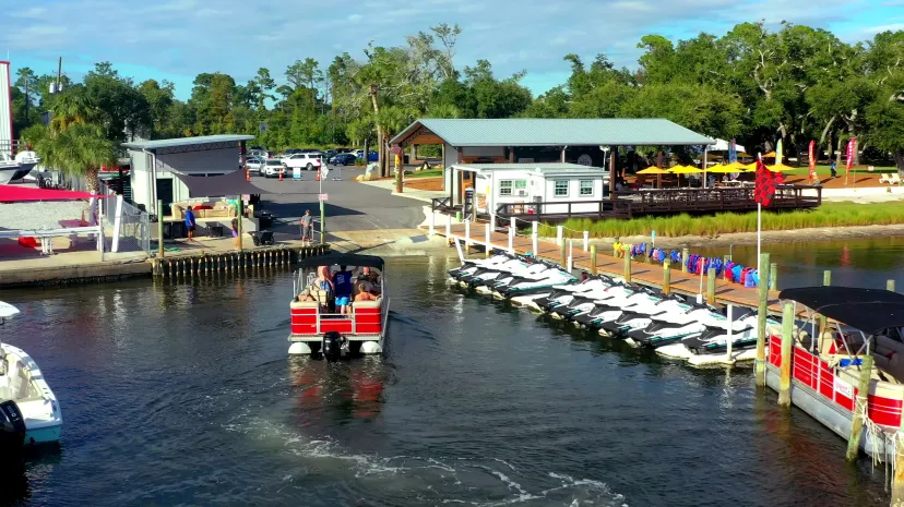 A small boat with passengers near a dock with parked boats and buildings.
