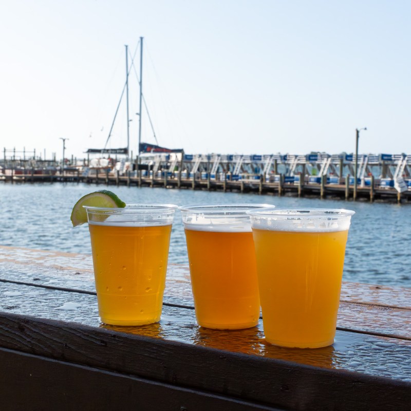 Three plastic cups of beer on a wooden table by a marina with boats.