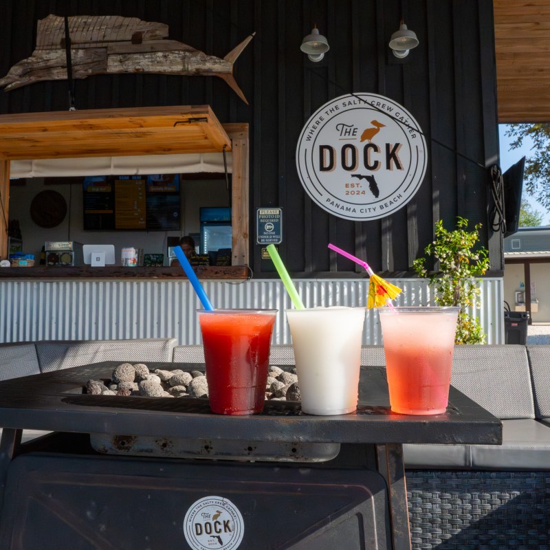 Three colorful drinks on a table at an outdoor bar with a 'The Dock' sign in the background.