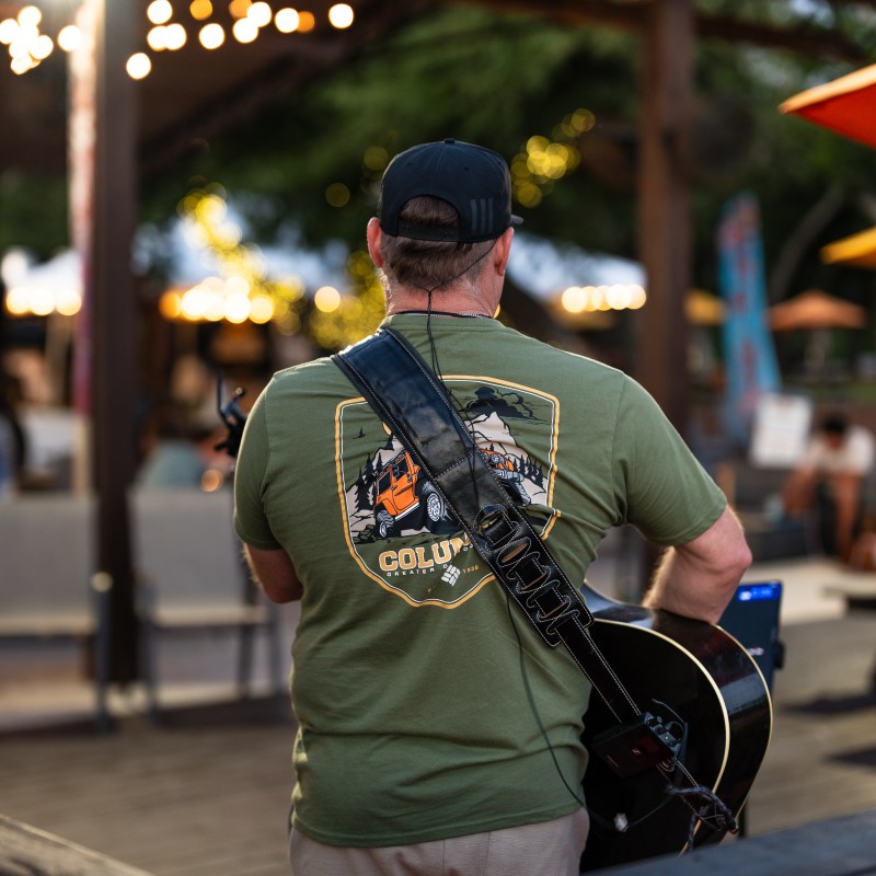 Musician with guitar performs outdoors, wearing a green shirt and black cap.