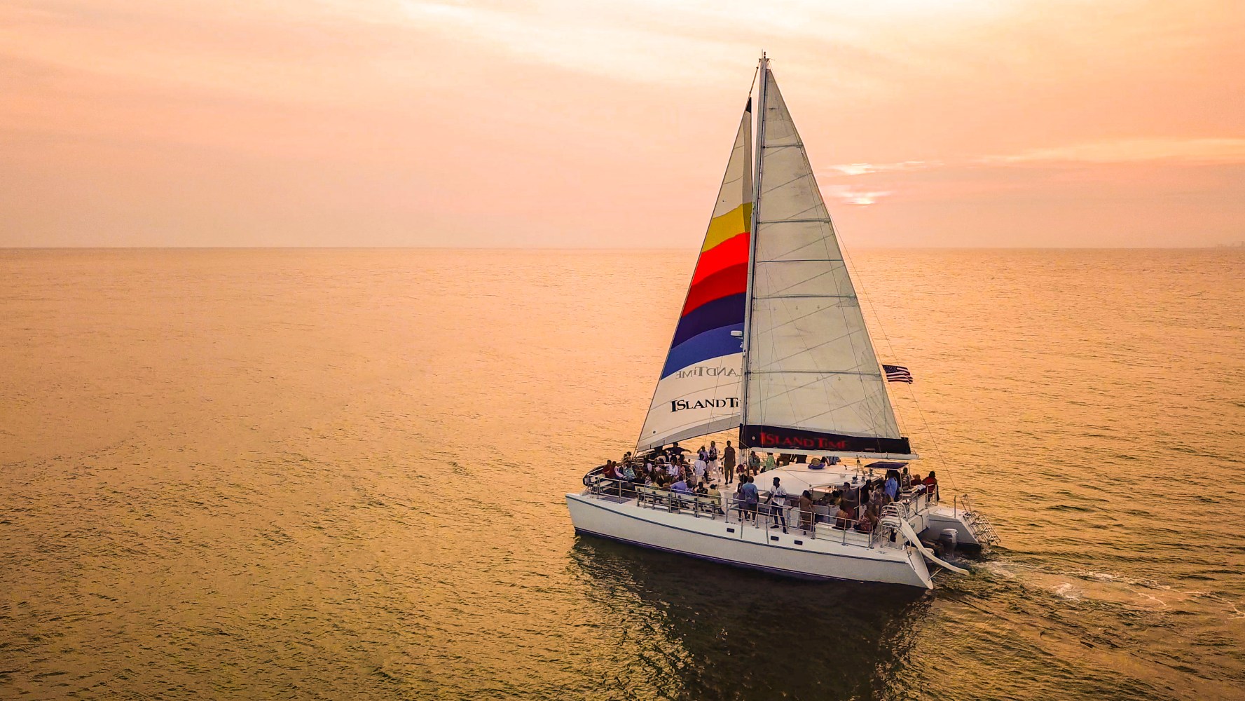 Sailboat with colorful sail on calm ocean at sunset, people on deck.
