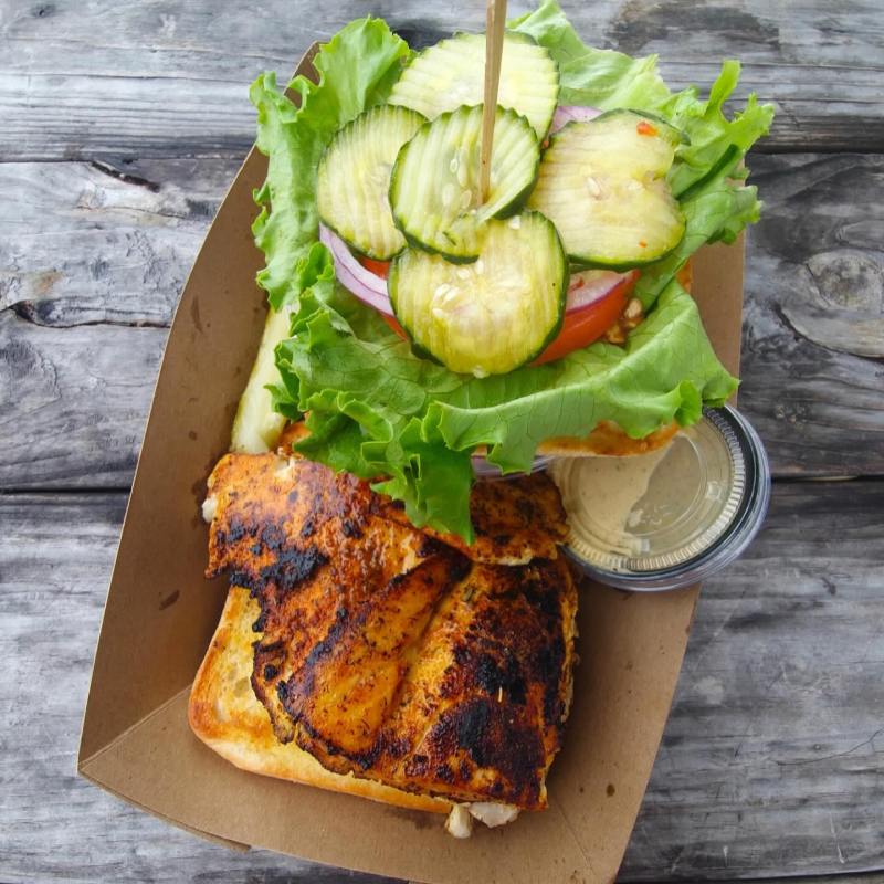 Grilled chicken sandwich with lettuce, pickles in a cardboard tray on wooden table.